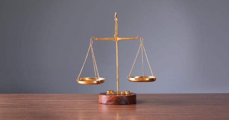 A small brass balance scale sits on a light wooden table, with a blurred laptop and documents in the background.