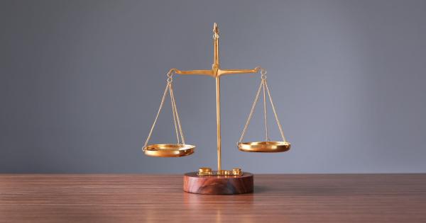 A small brass balance scale sits on a light wooden table, with a blurred laptop and documents in the background.
