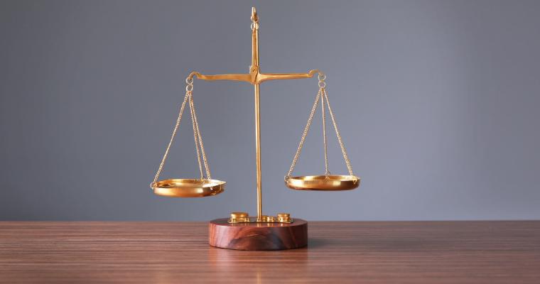 A small brass balance scale sits on a light wooden table, with a blurred laptop and documents in the background.